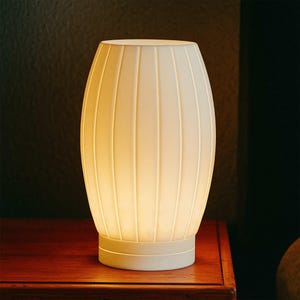 May include: A white, oval-shaped table lamp with vertical ribbed detailing. The lamp has a cylindrical base and a rounded top. The lamp is illuminated, casting a warm glow. The lamp is sitting on a wooden surface.