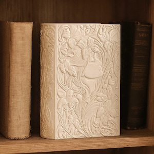 Könnte beinhalten: Ein cremefarbenes Buch mit einem geprägten Blumen- und Gesichtsdesign. Das Buch steht auf einem Holzregal neben zwei anderen Büchern. Das Cover zeigt ein detailliertes Relief eines Frauenprofils, umgeben von Blumen und Blättern.