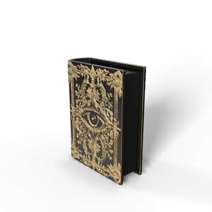 May include: Black book with gold filigree and an eye design on the cover. The ornate gold frame and eye are the focal point of the book's cover. The book is standing upright, showcasing its detailed design and craftsmanship.