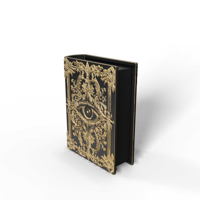 May include: Black book with gold filigree and an eye design on the cover. The ornate gold frame and eye are the focal point of the book's cover. The book is standing upright, showcasing its detailed design and craftsmanship.