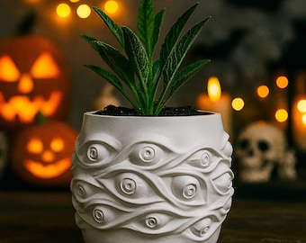 May include: A white ceramic plant pot featuring a repeating eye design, holding a green plant. The pot sits on a wooden surface, with blurred Halloween decorations in the background, including pumpkins and lights.