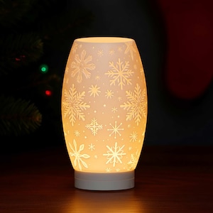 May include: A white, oval-shaped ceramic lamp with a snowflake design. The lamp is illuminated from within, casting a warm glow. The snowflake pattern is etched into the surface, creating a festive winter theme.