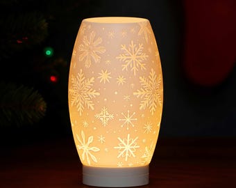 May include: A white, oval-shaped ceramic lamp with a snowflake design. The lamp is illuminated from within, casting a warm glow. The snowflake pattern is etched into the surface, creating a festive winter theme.