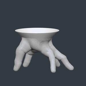 May include: A white, sculptural bowl supported by a pair of hands. The hands are detailed with visible veins and cracks, giving a realistic appearance. The bowl has a wide, shallow design, suitable for holding small items.