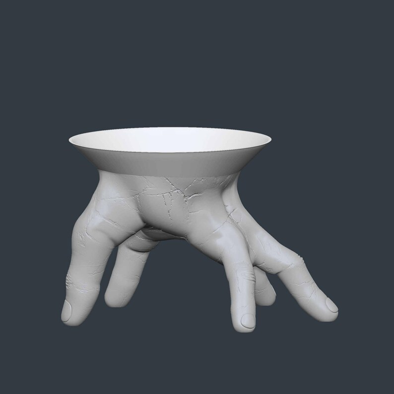 May include: A white, sculptural bowl supported by a pair of hands. The hands are detailed with visible veins and cracks, giving a realistic appearance. The bowl has a wide, shallow design, suitable for holding small items.