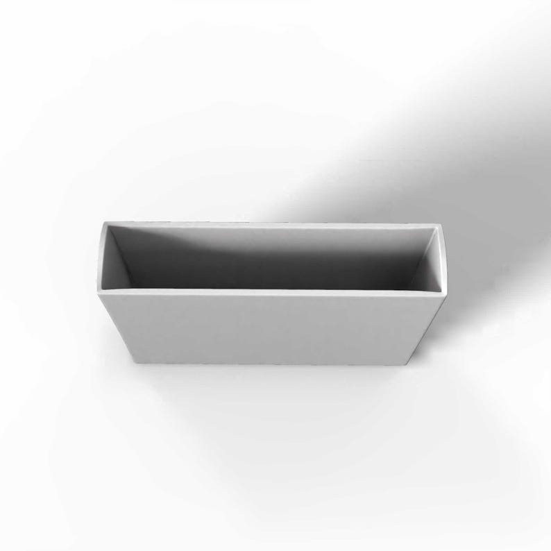 May include: A rectangular, light gray container with a slightly trapezoidal shape. The interior is a darker shade of gray, creating a contrast. The container is empty and sits against a white background.