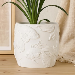 May include: White ceramic plant pot with an embossed koi carp design. The cylindrical pot features detailed relief carvings of two koi carp and flowing water elements. The pot holds a green plant with long, slender leaves, suitable for indoor or outdoor use.
