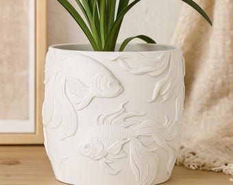 Puede incluir: Maceta de cerámica blanca con diseño de peces koi en relieve. La maceta cilíndrica presenta tallas en relieve detalladas de dos peces koi y elementos de agua. Contiene una planta verde con hojas largas y delgadas, apta para interior o exterior.