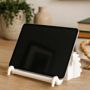 Puede incluir: Un soporte blanco para tablet con un diseño único, que sostiene una tablet negra. El soporte tiene una barra horizontal con brazos curvos que sostienen la tablet. El soporte está sobre una superficie de madera.
