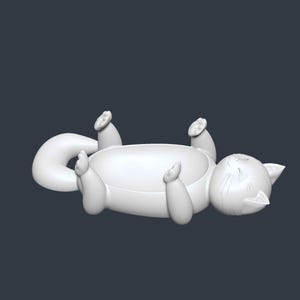 May include: A white cat-shaped decorative bowl, lying on its back with its paws and tail curved. The bowl has a rounded shape, with the cat's face and whiskers detailed on one end. The cat is in a relaxed pose.
