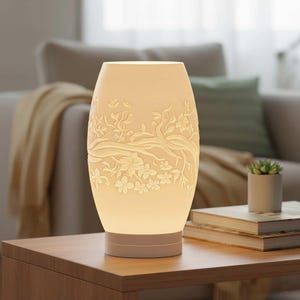 May include: A cream-colored, oval-shaped table lamp with a floral relief design. The lamp features a base and a top section that emits a soft glow. The floral pattern includes branches, leaves, and flowers. The lamp is on a wooden table.