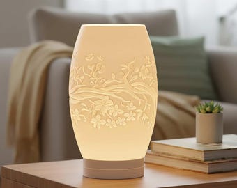 May include: A cream-colored, oval-shaped table lamp with a floral relief design. The lamp features a base and a top section that emits a soft glow. The floral pattern includes branches, leaves, and flowers. The lamp is on a wooden table.