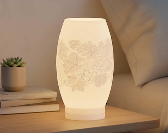 Könnte beinhalten: Eine leuchtende, weiße Keramiklampe mit einem Trauben- und Blätterdesign steht auf einem Nachttisch aus Holz. Die Lampe hat eine ovale Form und strahlt ein weiches Licht aus. Eine kleine Topf-Sukkulente und Bücher befinden sich ebenfalls auf dem Tisch.
