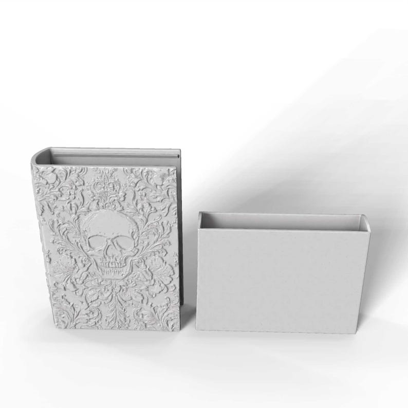 May include: A gray, book-shaped container with a skull and floral design, next to a rectangular container. The skull is centered on the front of the book, with ornate details. The containers are made of a solid material.