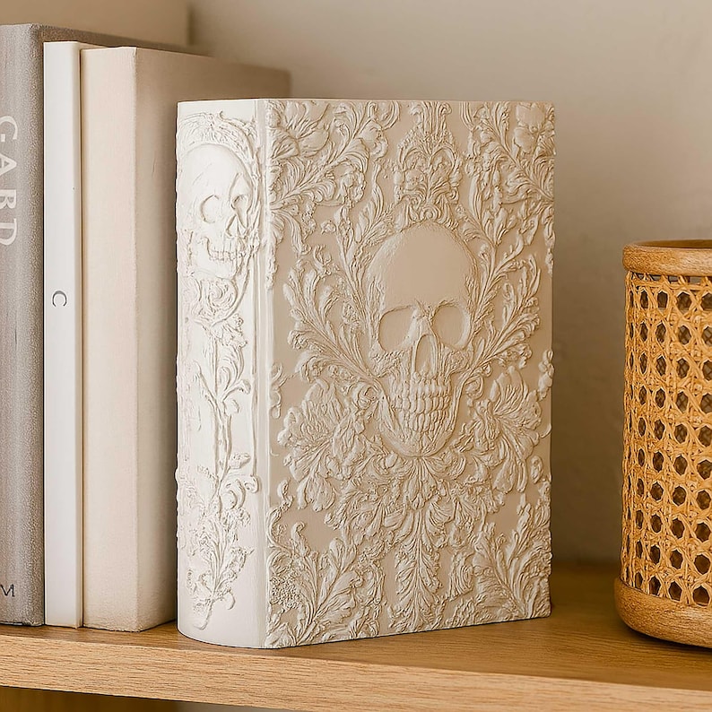 May include: Off-white decorative book with a skull design. The book has a detailed embossed pattern of skulls and floral elements. The spine features a skull, and the front cover showcases a larger skull surrounded by ornate foliage. The book is displayed on a wooden shelf.