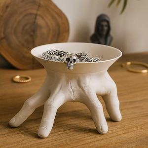 May include: A unique, off-white decorative bowl supported by a hand-shaped base. The bowl holds a silver chain and a skull-shaped charm. The bowl is placed on a wooden surface.