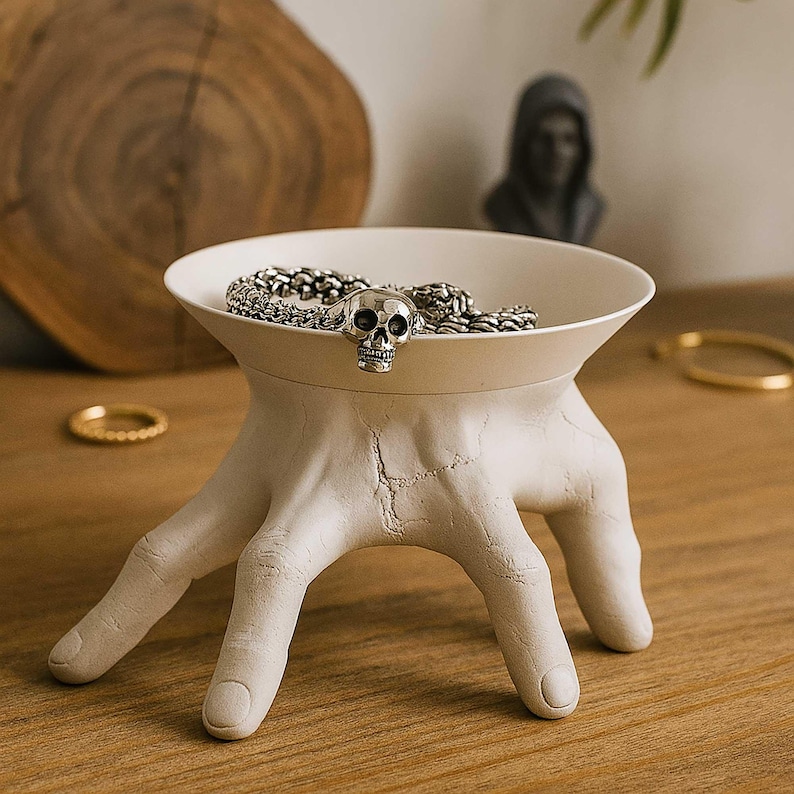 May include: A unique, off-white decorative bowl supported by a hand-shaped base. The bowl holds a silver chain and a skull-shaped charm. The bowl is placed on a wooden surface.