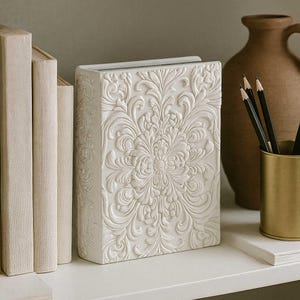 Puede incluir: Un libro decorativo blanco y rectangular con un diseño floral en relieve. El libro está de pie sobre una estantería blanca, junto a otros libros y un jarrón. Los lápices están en un soporte dorado.