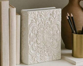 May include: A white, rectangular decorative book with an embossed floral design. The book is standing upright on a white shelf, next to other books and a vase. Pencils are in a gold-coloured holder.