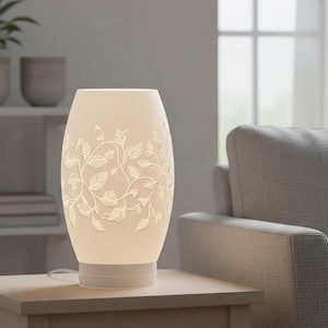 May include: A white, oval-shaped table lamp with a floral leaf design. The lamp has a cylindrical base and a soft, warm glow. The leaves and vines are etched into the lamp's surface, creating a decorative pattern. The lamp is on a light-colored table.