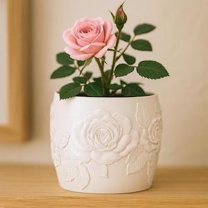 Pode incluir: Uma planta de rosas em vaso com uma flor rosa claro e folhas verdes. O vaso de cerâmica branca apresenta um desenho de rosa em relevo. A planta está sobre uma superfície de madeira.
