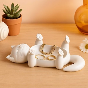 May include: A white cat-shaped jewelry tray with a raised paw design. The tray holds a gold chain bracelet and two gold rings. The cat is lying on its back, with its paws in the air. A succulent plant in a terracotta pot is in the background.