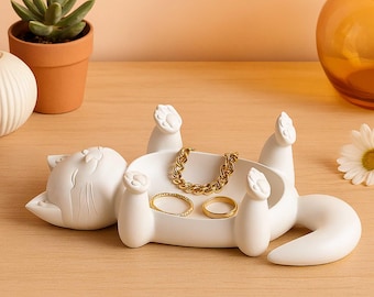 May include: A white cat-shaped jewelry tray with a raised paw design. The tray holds a gold chain bracelet and two gold rings. The cat is lying on its back, with its paws in the air. A succulent plant in a terracotta pot is in the background.