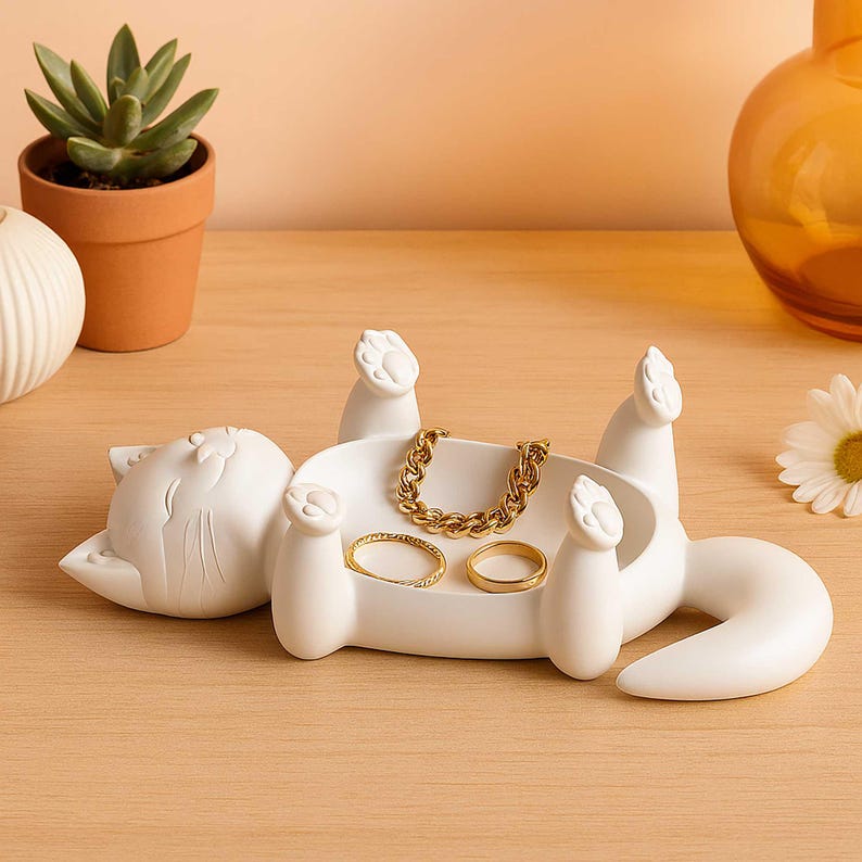 May include: A white cat-shaped jewelry tray with a raised paw design. The tray holds a gold chain bracelet and two gold rings. The cat is lying on its back, with its paws in the air. A succulent plant in a terracotta pot is in the background.