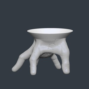 May include: A white, hand-shaped decorative bowl. The bowl's base is formed by a hand with fingers extended, supporting a circular, shallow dish. The surface has a textured, cracked appearance. The background is a solid, dark gray.