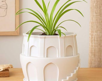 May include: A white ceramic planter with a unique architectural design, featuring arched openings and a staircase detail. The planter holds a vibrant green plant with long, slender leaves. The planter is approximately 8 inches in height.