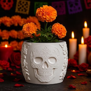 May include: A white ceramic planter with a skull design, holding vibrant orange marigold flowers. The planter is decorated with floral patterns and is surrounded by candles and flower petals, creating a Day of the Dead theme.