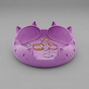 May include: A lavender cat-shaped jewelry dish with raised ears and etched facial features. The tray holds a collection of jewelry, including two gold-tone stud earrings, a gold band, a silver engagement ring, and two silver hoop rings.