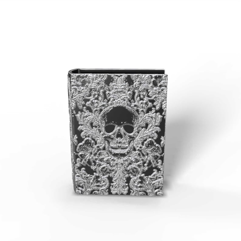 May include: A black and silver decorative book with a skull design. The cover features a detailed silver skull and ornate floral patterns. The book has a gothic or fantasy theme.