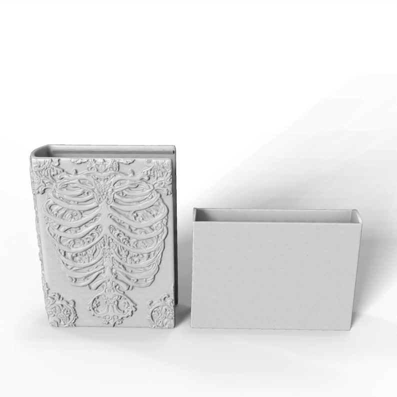 May include: A light gray, book-shaped container with a detailed rib cage design on the front cover. Beside it is a matching rectangular box. The container has an open top, suggesting it's a decorative storage item.