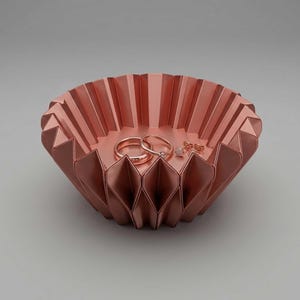May include: A metallic copper-colored bowl with a geometric, pleated, accordion-like structure. Inside the bowl, there are two plain metal rings and a pair of small stud earrings with tiny clear stones, all matching the copper tone of the bowl.