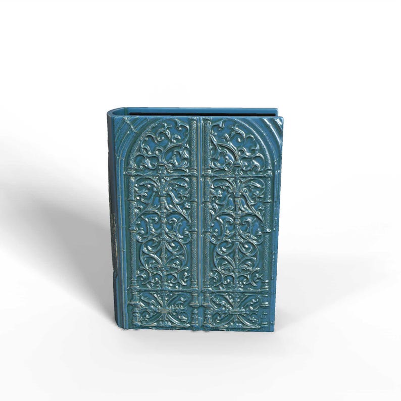 K&ouml;nnte beinhalten: Eine teal-farbene dekorative Box in Form eines Buches. Die Vorderseite zeigt verzierte, symmetrische T&uuml;rdesigns mit komplizierten Schn&ouml;rkeln. Die Box hat eine leicht verwitterte Oberfl&auml;che, die ihr ein Vintage-Aussehen verleiht.