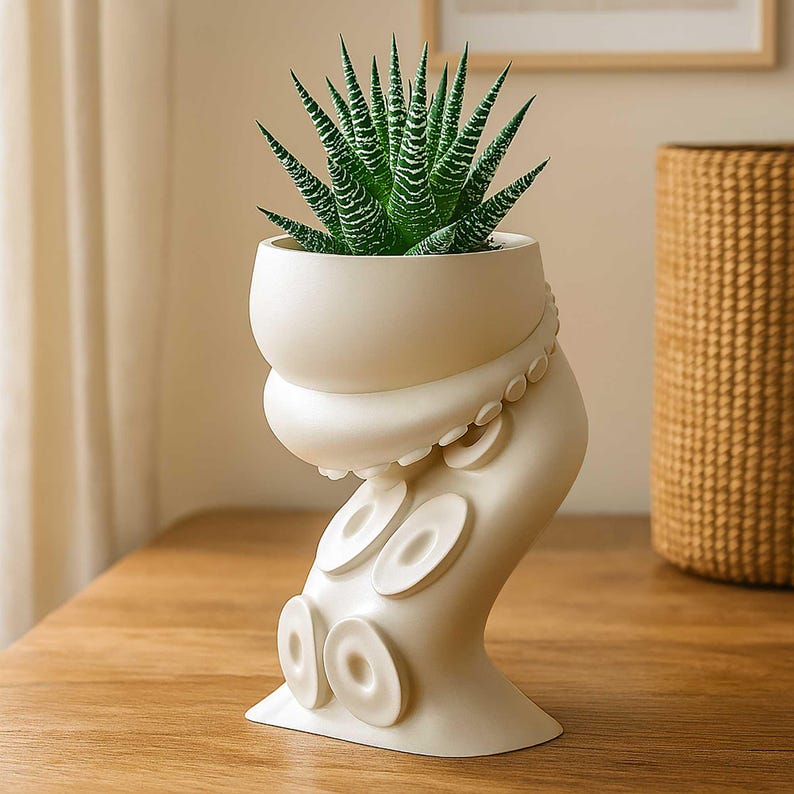 May include: A cream-colored ceramic planter shaped like an octopus tentacle, holding a green and white succulent plant. The planter has a rounded top and a base with circular details. The plant has pointed, striped leaves. The planter is on a wooden surface.