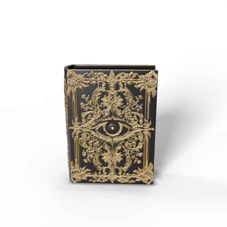 May include: A black book with gold filigree and an eye design. The cover features ornate gold detailing, including floral patterns and an eye symbol. The book has a rectangular shape and a slightly curved spine.