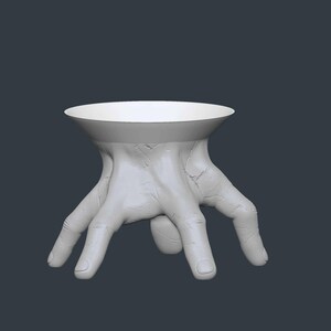 May include: A white, sculptural table with a unique design. The table's base is formed by a hand with fingers spread out, supporting a circular tabletop. The overall aesthetic is modern and artistic, suitable for decorative purposes.