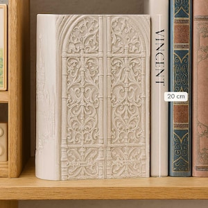 May include: Off-white bookend featuring a detailed relief of ornate double doors. The bookend is next to a book with the name "VINCENT" and a label indicating a height of 20 cm, approximately 7.87 inches. The bookend is on a wooden shelf.