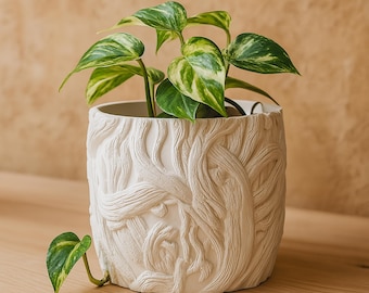 Puede incluir: Una maceta de cerámica blanca con un diseño detallado de corteza de árbol, que contiene una planta de potos verde y amarilla. La planta tiene hojas en forma de corazón. La maceta está sobre una superficie de madera.