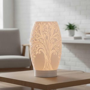 May include: A white ceramic table lamp with a tree of life design. The lamp has an oval shape and sits on a white base. The tree design is etched into the lamp's surface, creating a detailed pattern. The lamp is on a wooden table.