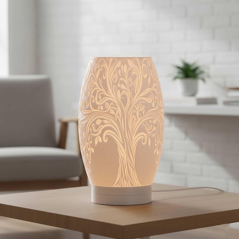 May include: A white ceramic table lamp with a tree of life design. The lamp has an oval shape and sits on a white base. The tree design is etched into the lamp's surface, creating a detailed pattern. The lamp is on a wooden table.