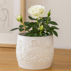 Pode incluir: Uma planta de rosas brancas num vaso de cerâmica branco com detalhes florais em relevo. O vaso tem formato cilíndrico e assenta sobre uma superfície de madeira. A rosa tem uma flor totalmente desabrochada, botões e folhas verdes.