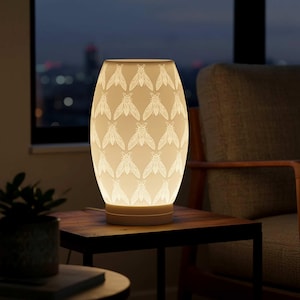 May include: A cream-colored, oval-shaped table lamp with a repeating pattern of white insect designs. The lamp is illuminated, casting a warm glow. It sits on a dark wooden table, with a chair and a window in the background.