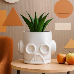 May include: A white ceramic planter shaped like a helmet, with two circular eye openings and a rectangular mouth. The planter holds a green plant with long, pointed leaves. Two apples sit on a wooden table next to the planter.
