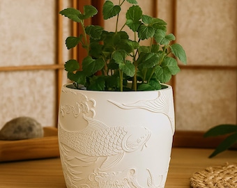 Puede incluir: Una maceta de cerámica blanca con un relieve detallado de una carpa koi y olas. La maceta está llena de plantas verdes de hojas. El diseño es un motivo clásico de inspiración asiática. Un artículo decorativo.