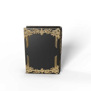 May include: A black book with gold decorative trim. The trim features ornate floral and scroll designs. The book's spine is curved, and the gold trim is on the front, back, and spine. The book is a decorative item.