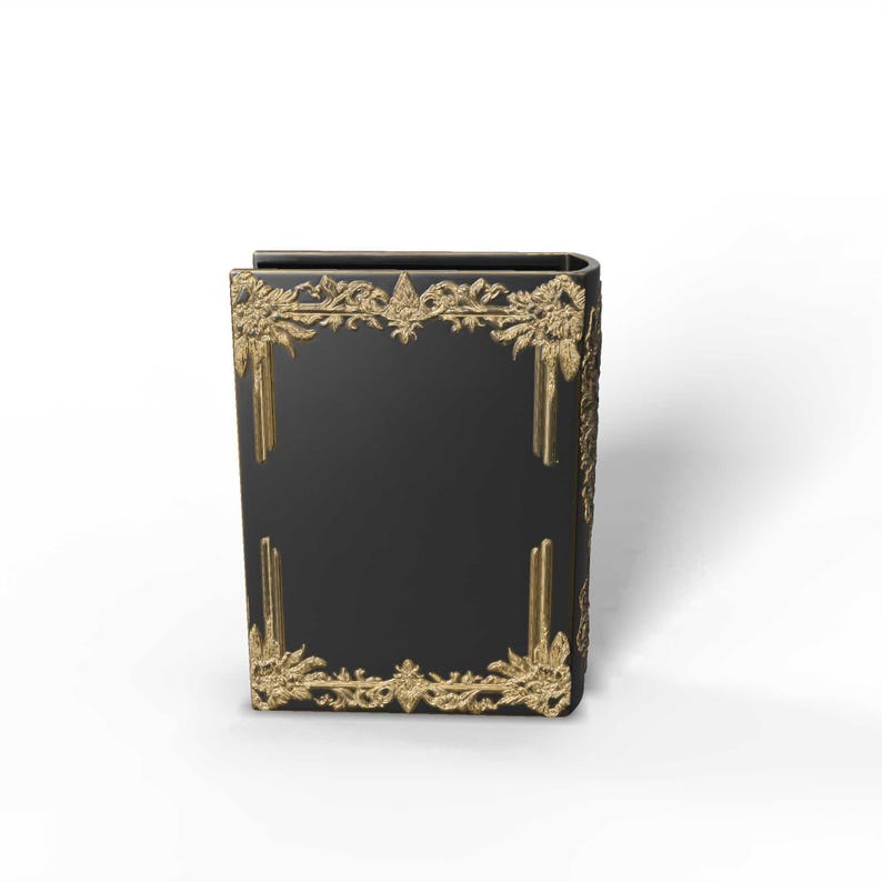 May include: A black book with gold decorative trim. The trim features ornate floral and scroll designs. The book's spine is curved, and the gold trim is on the front, back, and spine. The book is a decorative item.