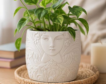 May include: A white ceramic plant pot featuring a detailed relief of a woman's face and floral patterns. The pot contains a green plant with patterned leaves. It rests on a round, woven tray, suitable for home decoration.
